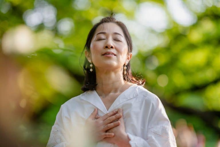 Nasal Obstruction Patient Needs Latera Woman in a park. Upper body and head are in frame. Her hands are clasping her chest. Her head is tilted slightly back and her mouth is barely open. Her eyes are closed in a look of joyful contemplation. The background is very blurry but includes the shape of trees and people.